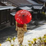 Seorang warga Jepang berjalan di permukiman tradisional dengan payung merah, ilustrasi kehidupan sehari-hari di tengah wacana penghapusan pajak makanan di Jepang